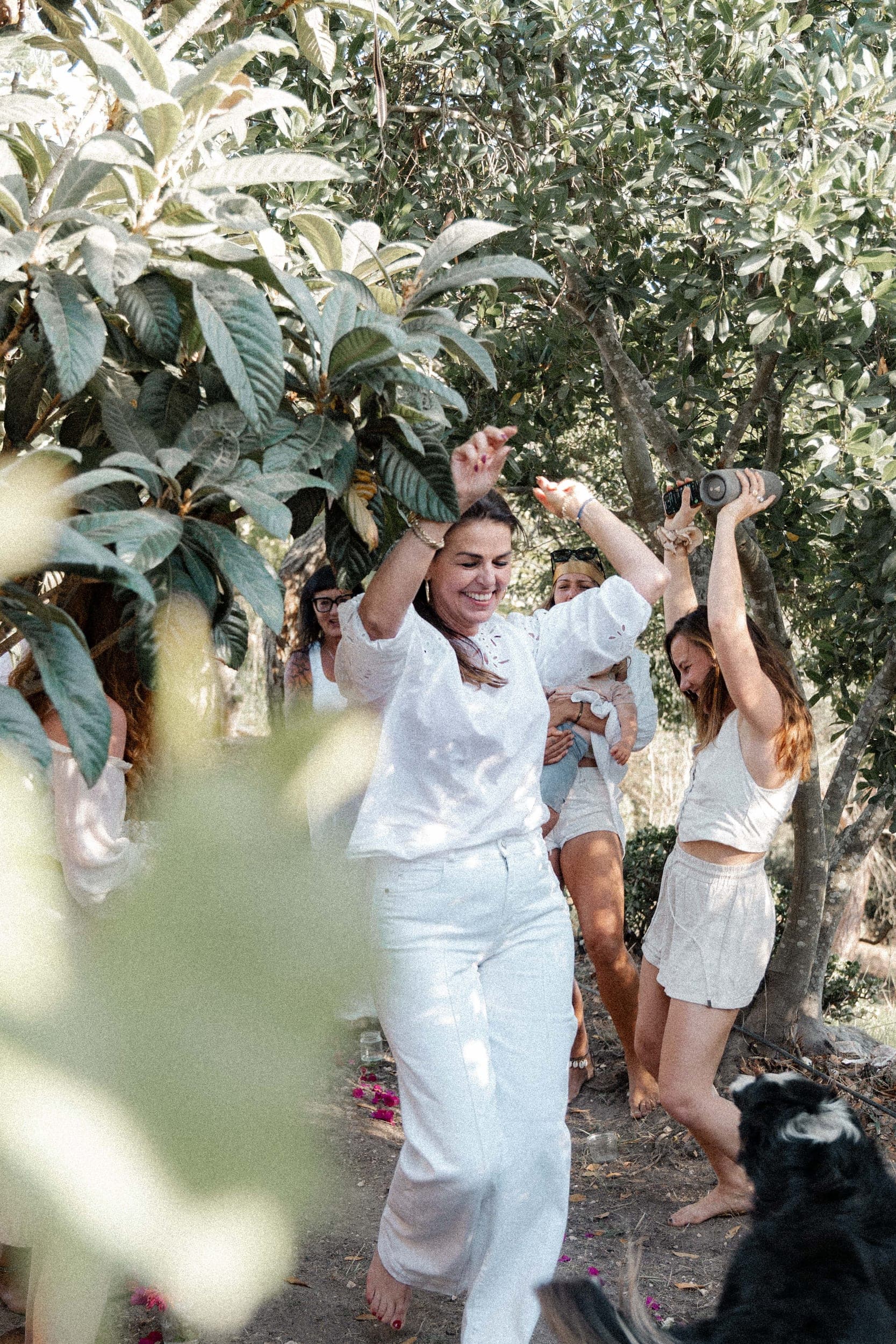 Women in white dancing joyfully under leafy trees with a dog during an outdoor retreat